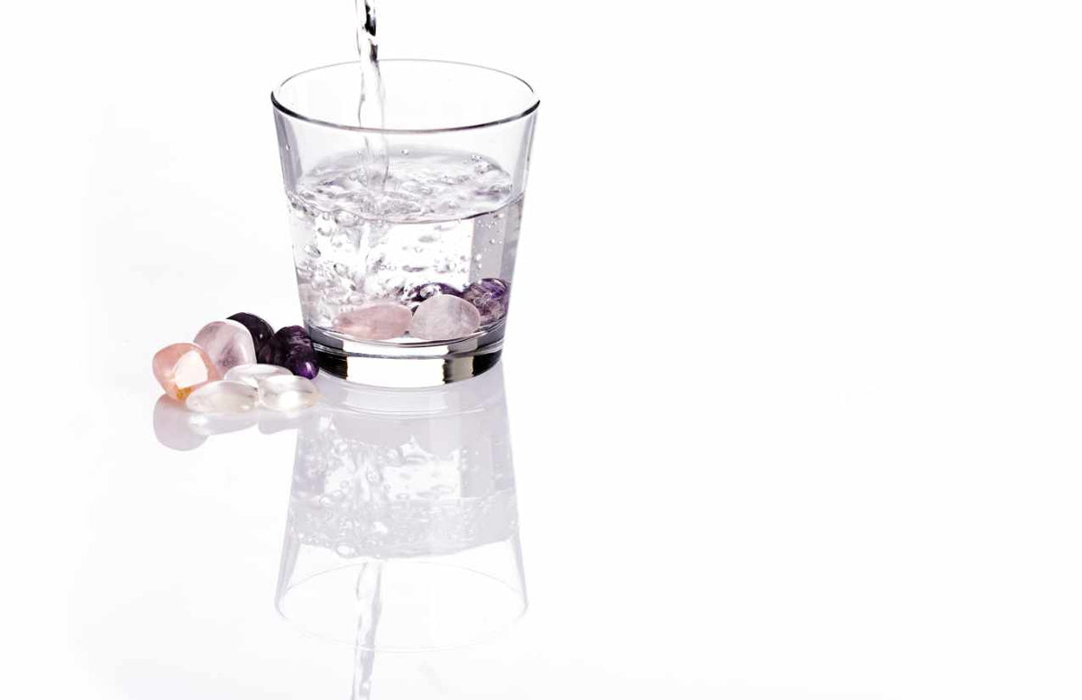 Wasser fließt in ein Glas mit Edelsteinen, weitere Trommelsteine liegen daneben auf weißem Grund