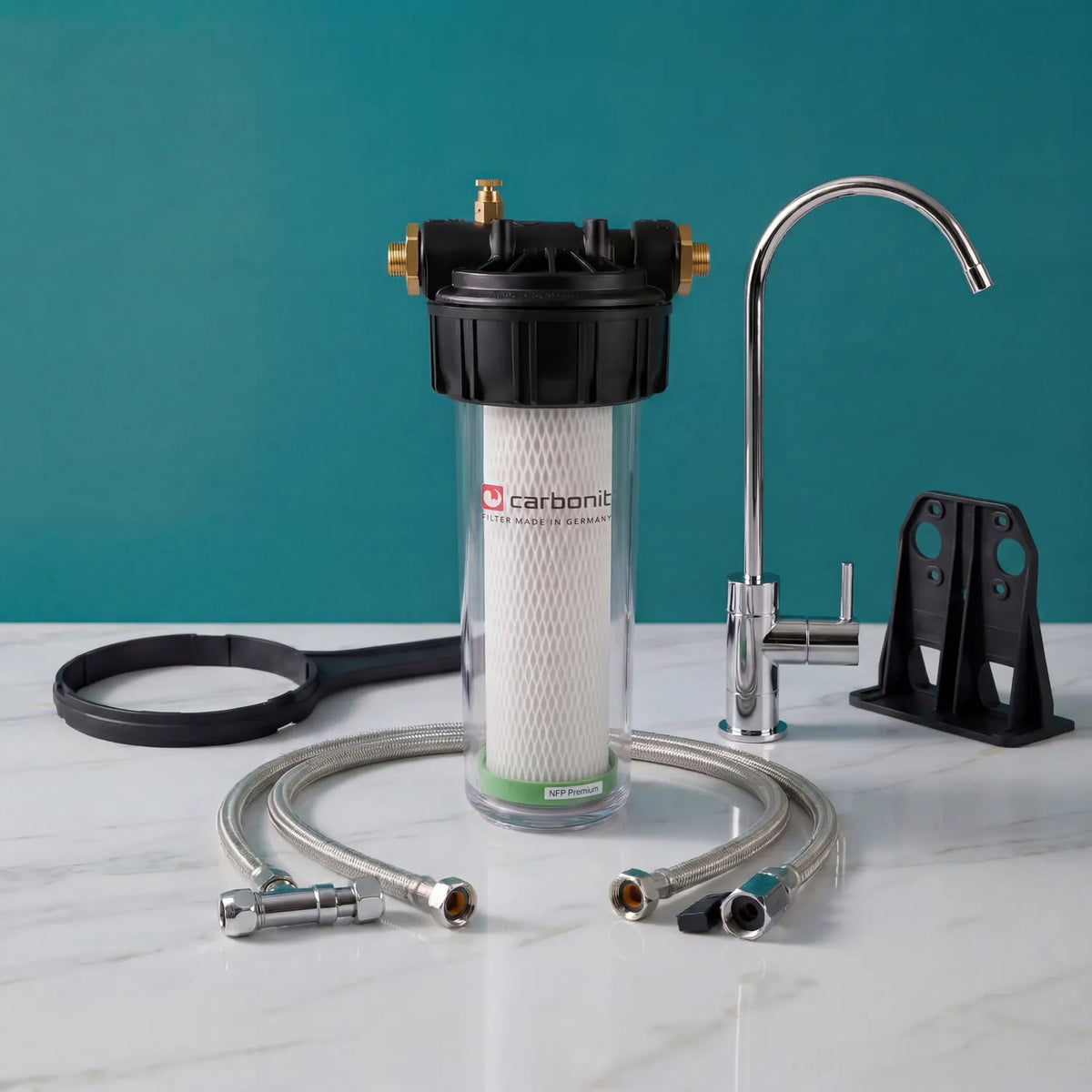 Carbonit Vario Classic Einbaufilter-Set auf weißer Marmorarbeitsplatte – transparentes Gehäuse mit NFP-Premium-Filterkartusche (Kreuzstruktur, grüner Ring), schwarzer Kopf mit Messing-Gewindeanschlüssen und Entlüftungsschraube, schwarzer Ringschlüssel und Wandhalterung, Beistell-Trinkhahn (Schwanenhals mit Hebel), zwei Schläuche; teal Hintergrund