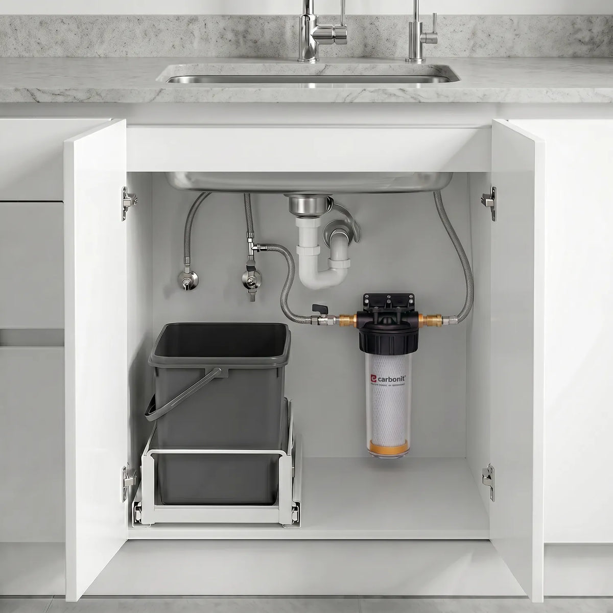 Carbonit Vario-HP Comfort im offenen Unterschrank – transparentes Gehäuse mit IFP-Puro-Kartusche (orange Bodenkappe) und schwarzem Kopf, rechts an weißer Schrankwand montiert; grauer Müllcontainer auf Ausziehschienen links; Edelstahlspüle, P-Trap, Absperrventile und Zuleitungen oben; Marmor-Arbeitsplatte und 1-Wege-Hahn, helle moderne Küche