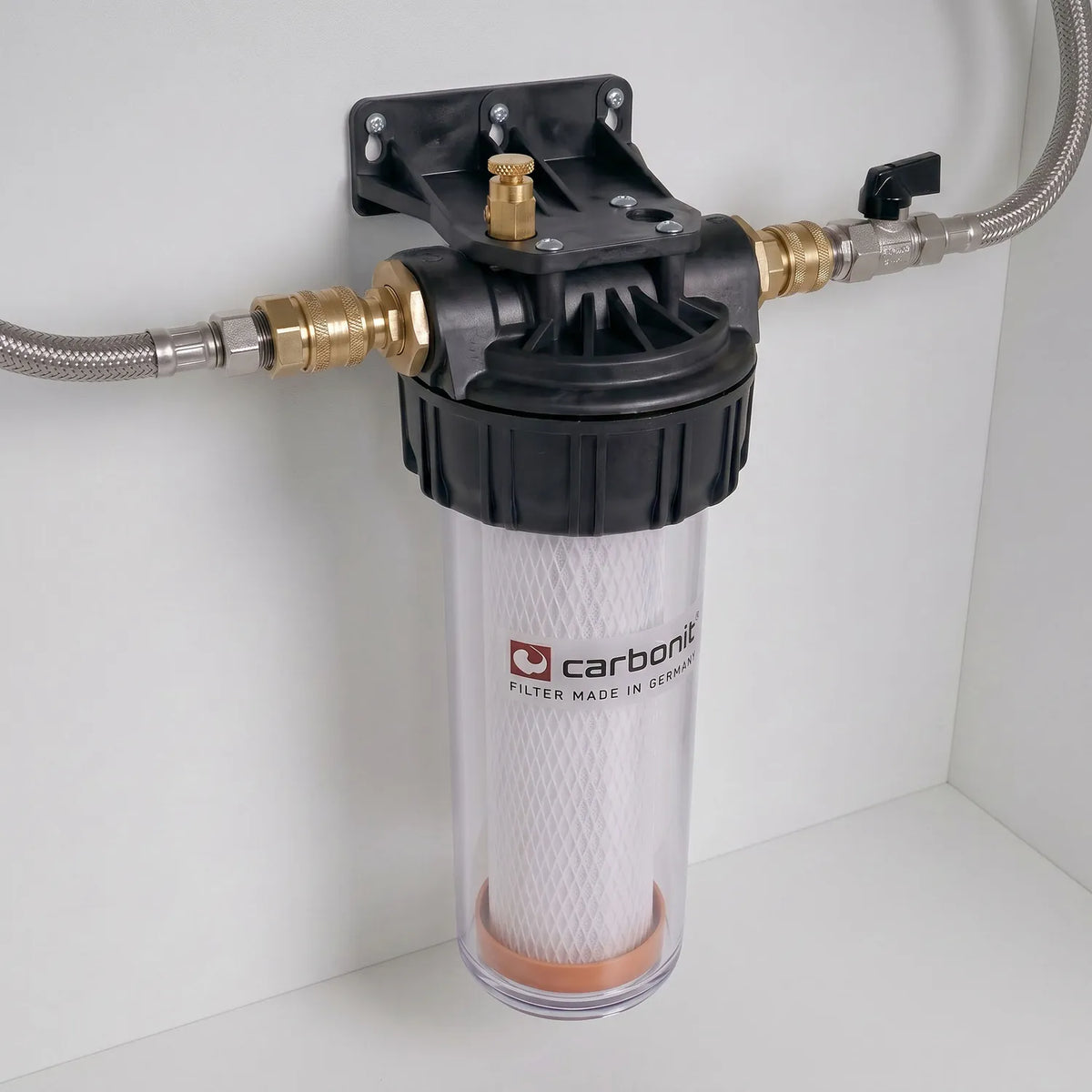 Carbonit Vario-HP Universal montiert an weißer Schrankwand – transparentes Gehäuse mit weißer IFP-Puro-Kartusche (orangene Bodenkappe), schwarzer Kopf mit Messing-Entlüftungsschraube, Schläuche links und rechts (rechts mit schwarzem Kugelventil-Hebel)