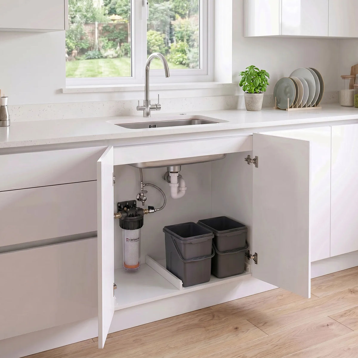 Carbonit Vario-HP Universal im offenen Unterschrank einer hellen weißen Küche – transparentes Gehäuse mit Kartusche und schwarzem Kopf, links an Schrankwand montiert; 3-Wege-Hahn, Gartenfenster, Basilikum auf der Arbeitsplatte
