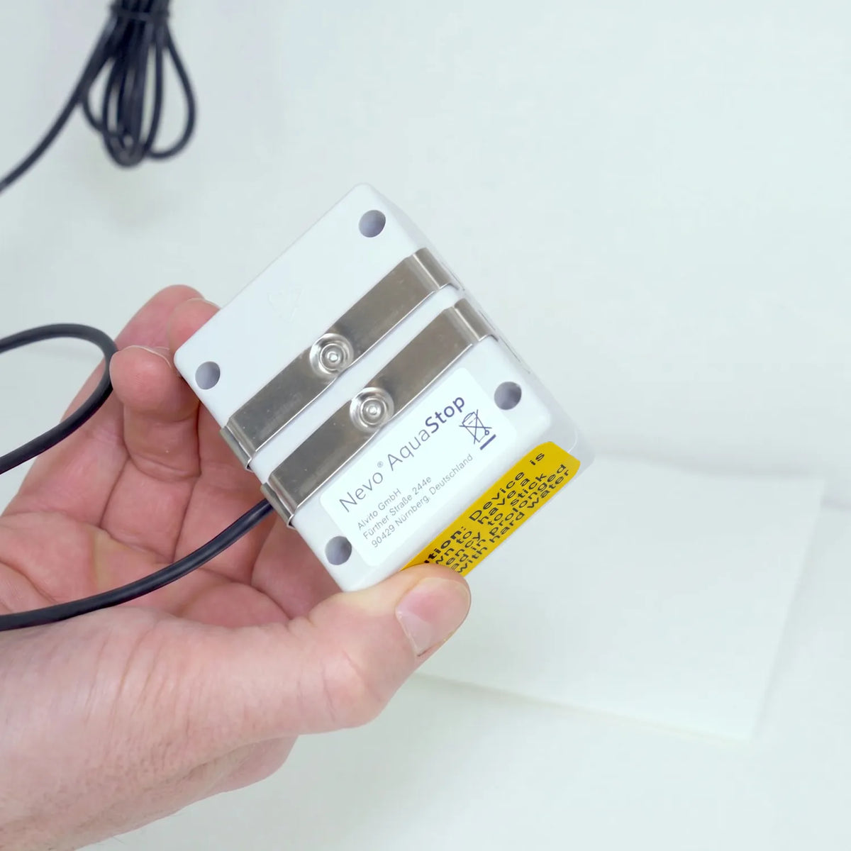Hand hält Nevo AquaStop AS-413 Controller mit Unterseite zur Kamera – Edelstahl-Sensorkontakte, weißes Typenschild mit Aufschrift „Nevo AquaStop, Alvito GmbH, Fürther Straße 244e, 90429 Nürnberg Deutschland", gelbes CE-Etikett; weißer Hintergrund
