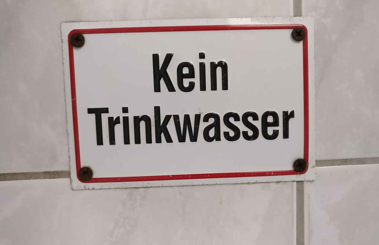 Weißes Emailleschild mit rotem Rand und der Aufschrift „Kein Trinkwasser“ an einer gefliesten Wand