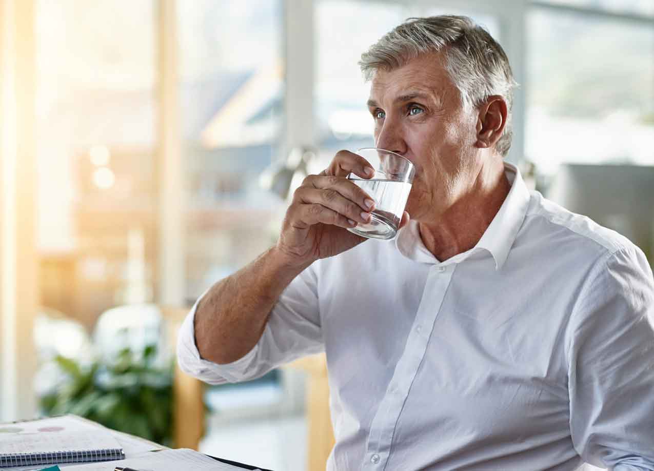 Älterer Mann im weißen Hemd trinkt am Schreibtisch ein Glas Wasser