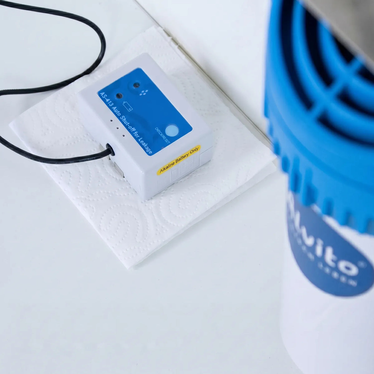 Nevo AquaStop AS-413 Controller von oben auf weißer Küchenrolle – weißes Kunststoffgehäuse mit blauem Bedienfeld und schwarzem Kabel; im unscharfen rechten Bildhintergrund blauer Deckel des Alvito Einbaufilters mit Alvito-Logo sichtbar