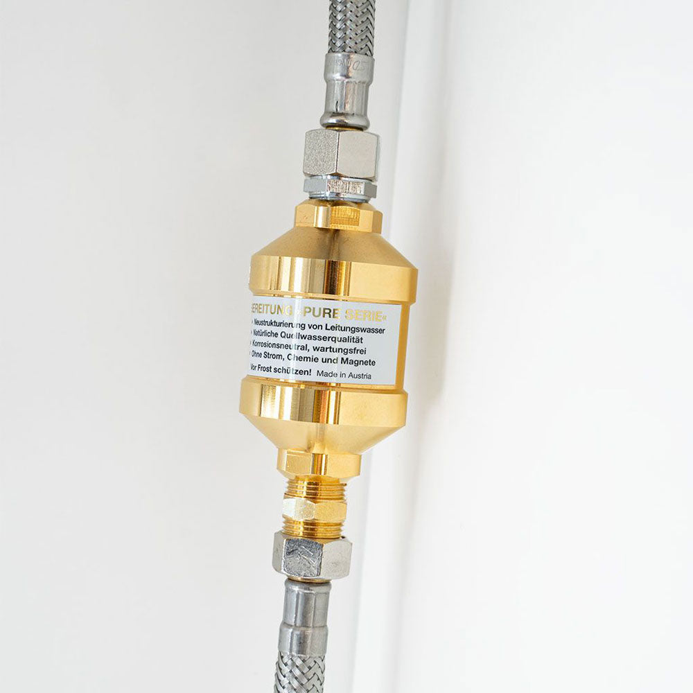 Goldener Inline-Wasseraufbereiter der Pure Serie zwischen zwei Edelstahlschläuchen an weißer Wand installiert